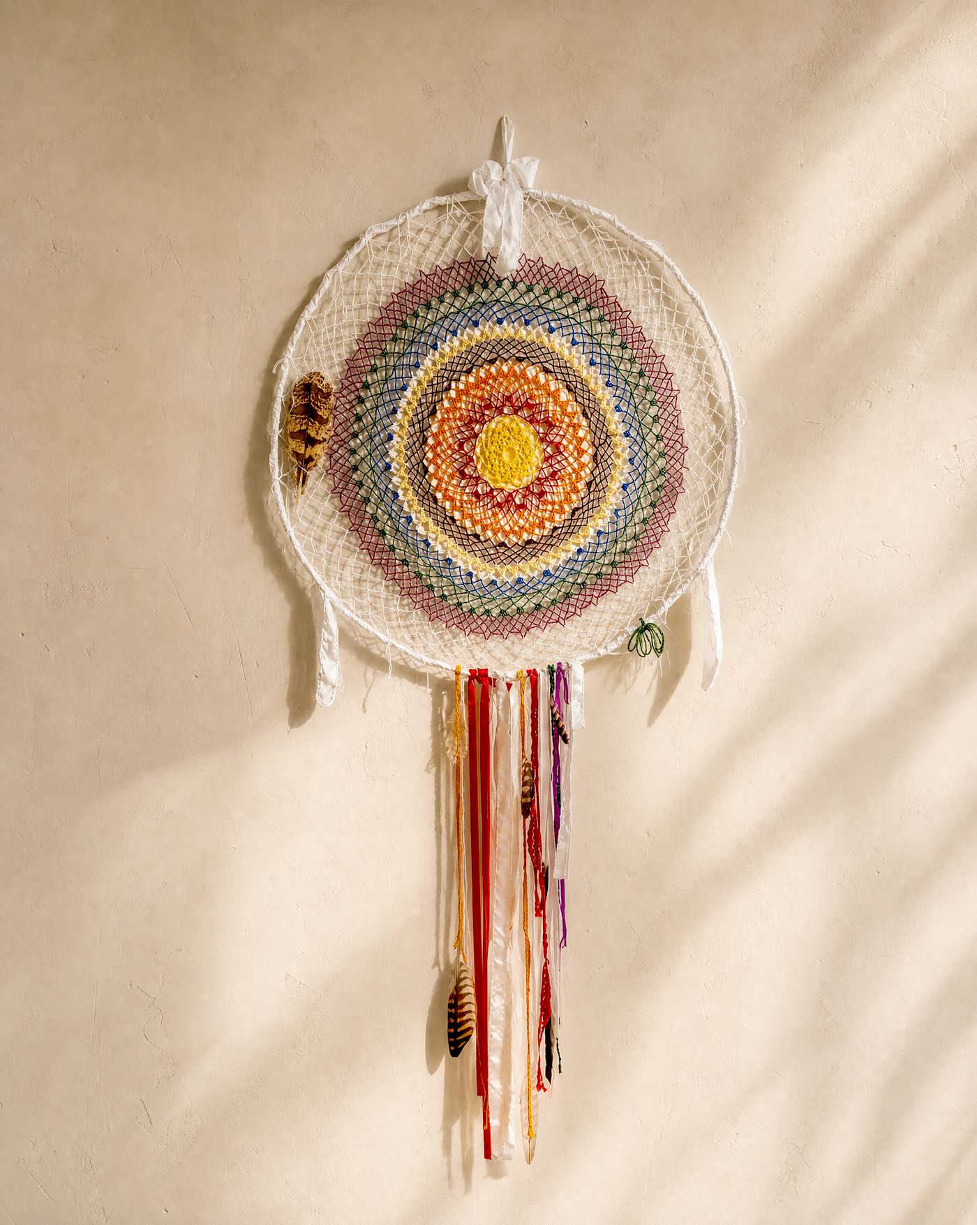 The Watcher, a handcrafted dreamcatcher with a concentric multicoloured weave, hung against a plain wall.